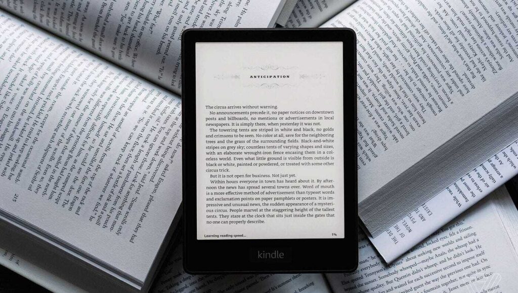 A black Amazon Kindle e-reader resting directly on top of an open physical book. The Kindle screen displays the chapter title "ANTICIPATION" and begins with the text, "The circus arrives without warning.